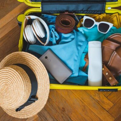 Open yellow suitcase packed with vacation items including straw hat, sunglasses, sandals, headphones, phone, and blue clothing on wooden floor