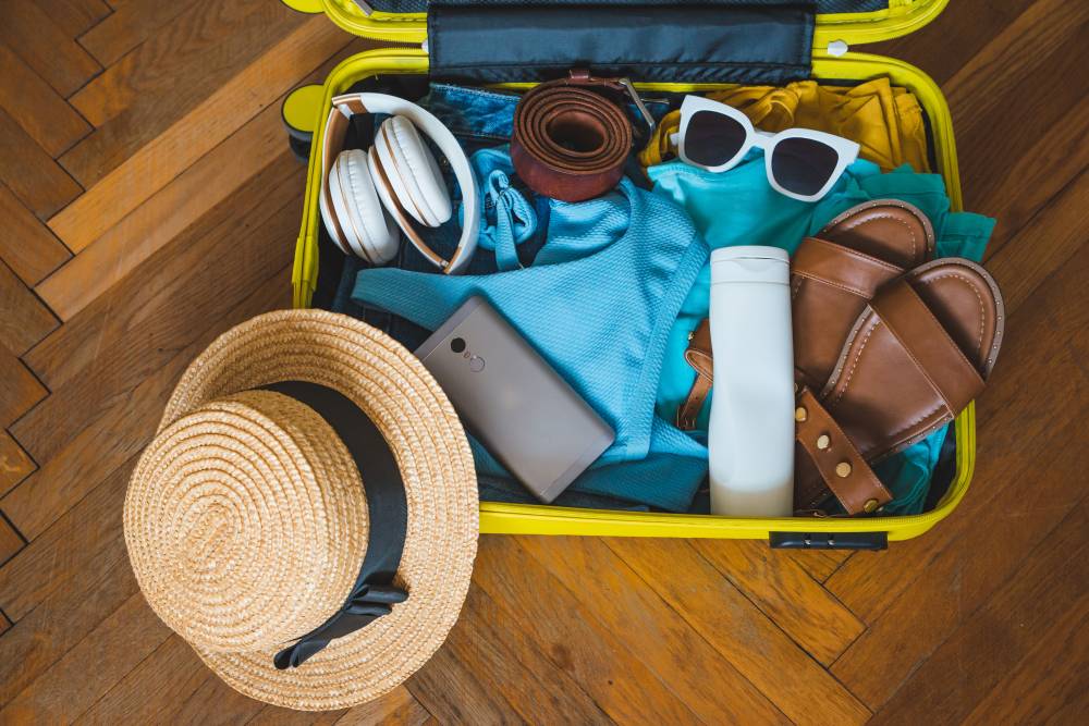 Open yellow suitcase packed with vacation items including straw hat, sunglasses, sandals, headphones, phone, and blue clothing on wooden floor