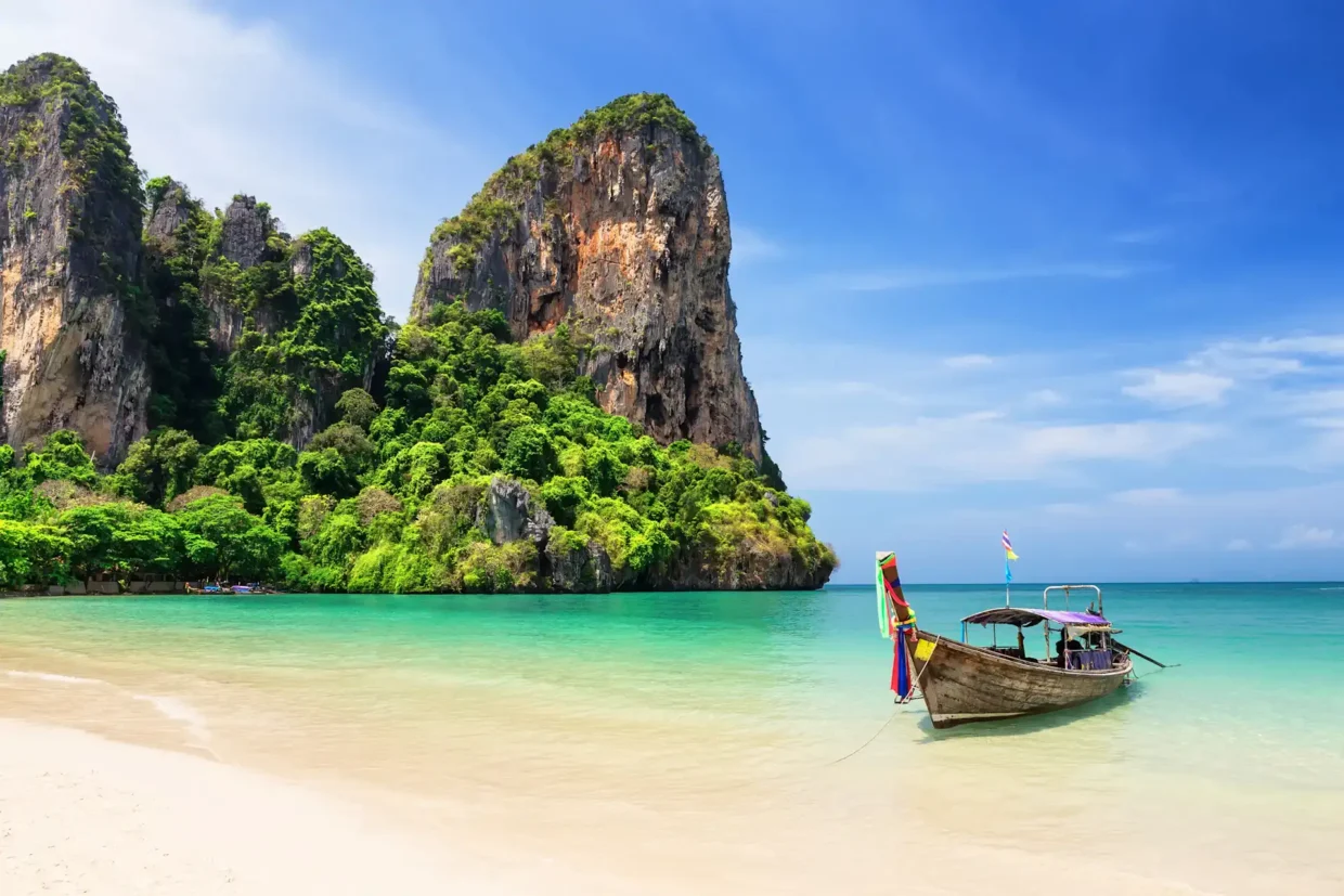 Traditional longtail boat on turquoise waters at Thai beach with dramatic limestone cliffs and lush tropical vegetation