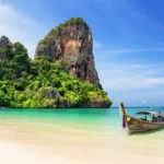 Traditional longtail boat on turquoise waters at Thai beach with dramatic limestone cliffs and lush tropical vegetation