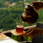 Person pouring tea from traditional clay teapot into glass cup on wooden tray outdoors with green foliage background