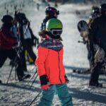 Child skier in green helmet and red jacket on snowy slope surrounded by other skiers at mountain resort