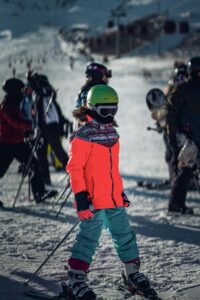 Child skier in green helmet and red jacket on snowy slope surrounded by other skiers at mountain resort