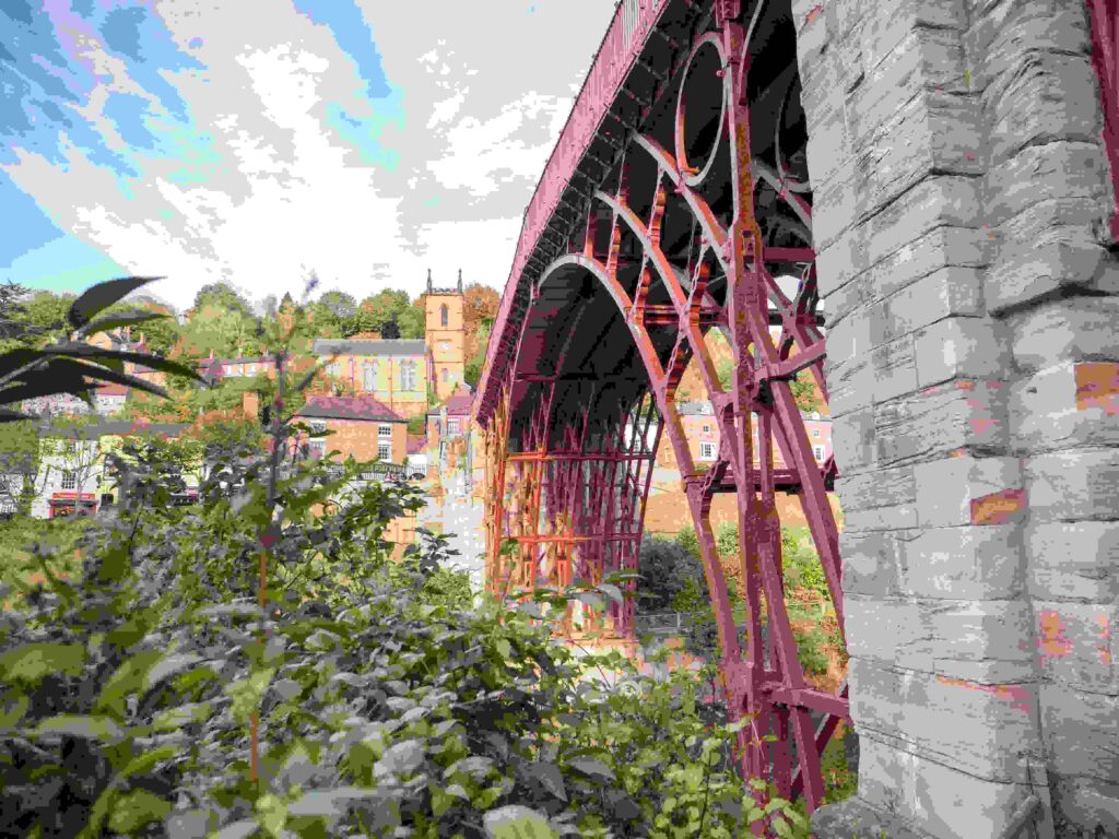Historic Iron Bridge with red metal framework spanning valley, surrounded by lush greenery and traditional buildings