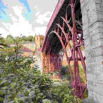 Historic Iron Bridge with red metal framework spanning valley, surrounded by lush greenery and traditional buildings