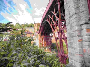 Historic Iron Bridge with red metal framework spanning valley, surrounded by lush greenery and traditional buildings