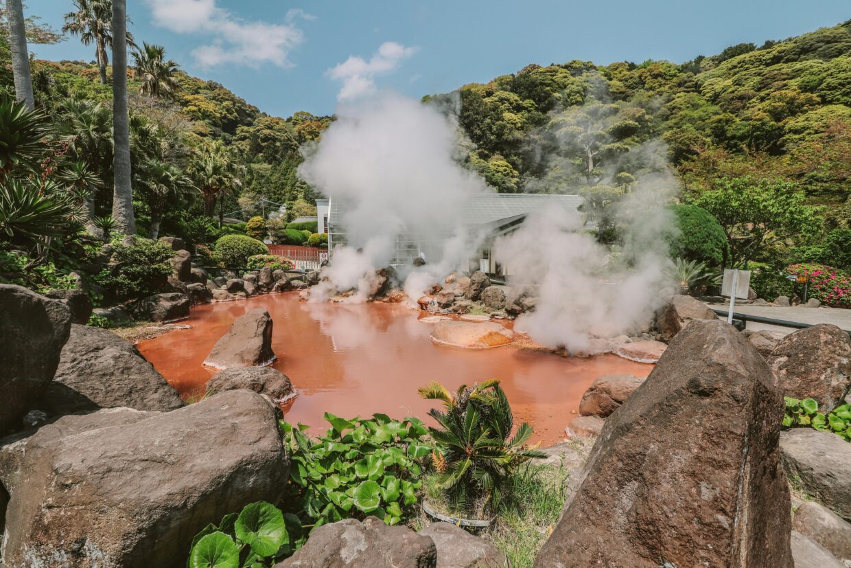 Red-orange hot spring with steam rising, surrounded by volcanic rocks and lush tropical vegetation in a mountainous setting.