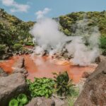 Red-orange hot spring with steam rising, surrounded by volcanic rocks and lush tropical vegetation in a mountainous setting.
