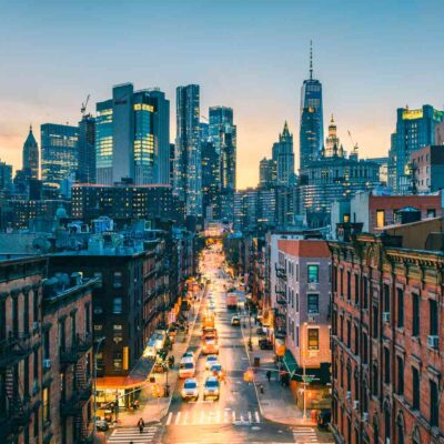 New York City street scene at dusk with Manhattan skyline, brick buildings, traffic, and One World Trade Center visible