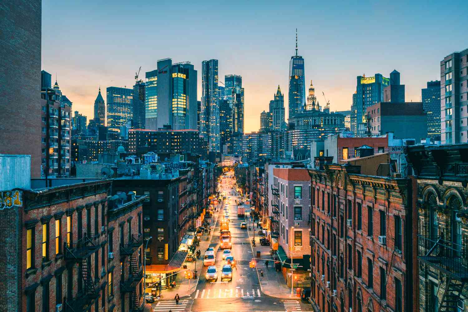 New York City street scene at dusk with Manhattan skyline, brick buildings, traffic, and One World Trade Center visible