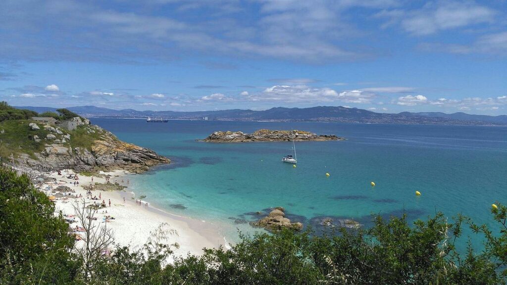 Pristine white sand beach with turquoise waters, sailboat, rocky outcrops, and lush green vegetation on Cíes Islands