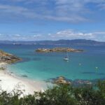 Pristine white sand beach with turquoise waters, sailboat, rocky outcrops, and lush green vegetation on Cíes Islands