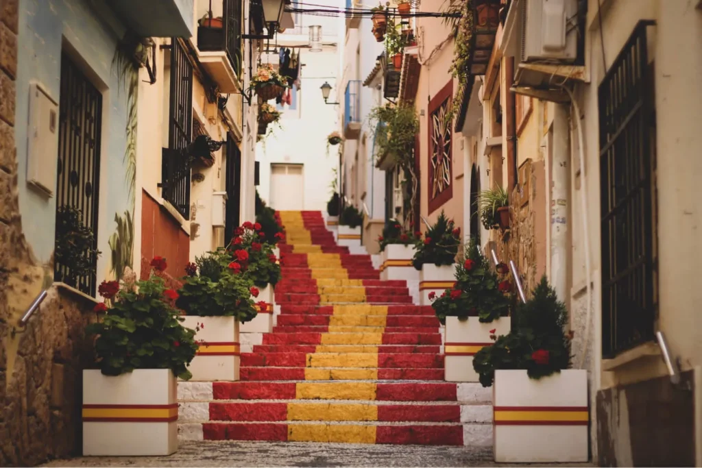 Colorful painted stone steps in red and yellow stripes leading up a narrow Mediterranean street lined with flowers and balconies.