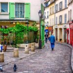 Charming cobblestone street in European village with colorful buildings, green shutters, outdoor cafe, and person walking