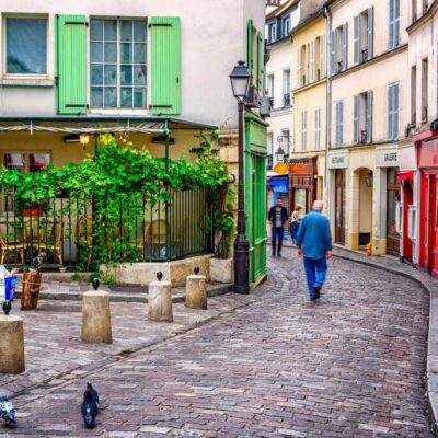 Charming cobblestone street in European village with colorful buildings, green shutters, outdoor cafe, and person walking
