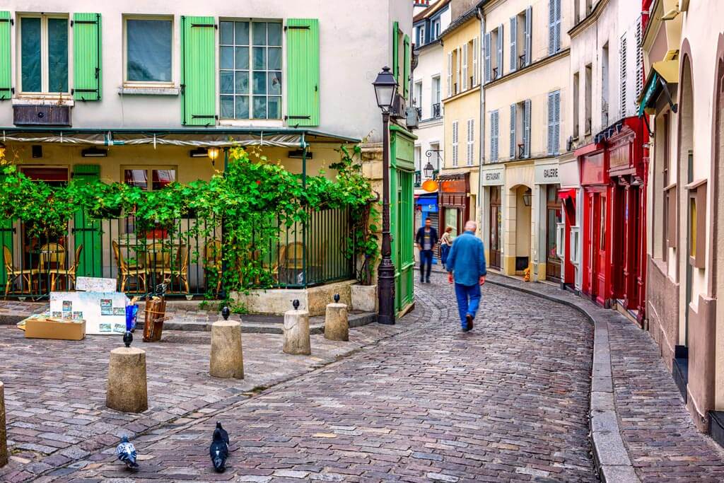 Charming cobblestone street in European village with colorful buildings, green shutters, outdoor cafe, and person walking