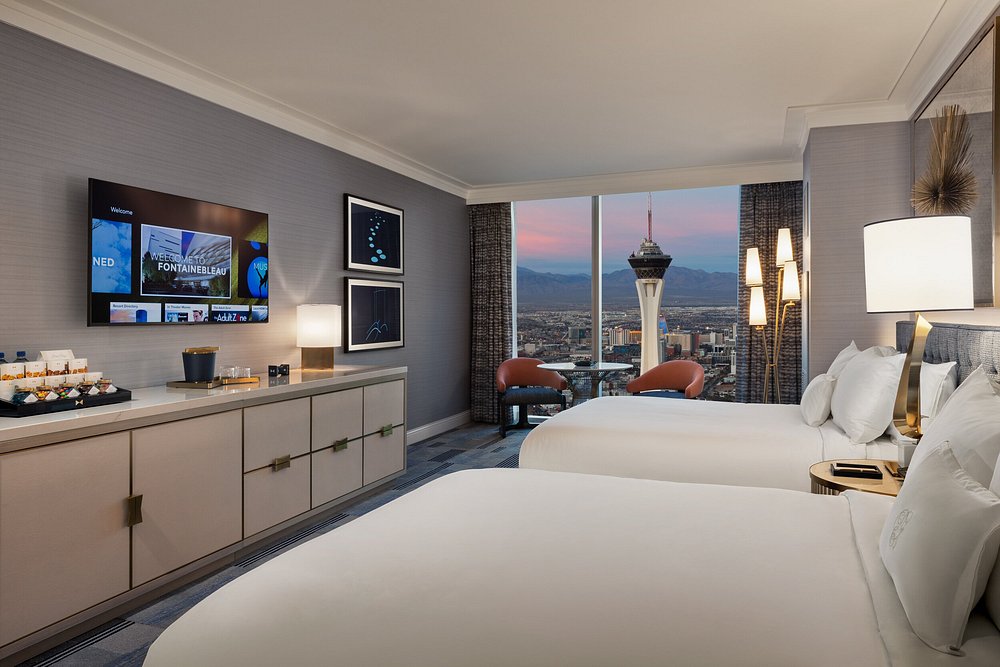 Luxury hotel room with two beds, floor-to-ceiling windows showing Las Vegas skyline, modern furniture and large TV