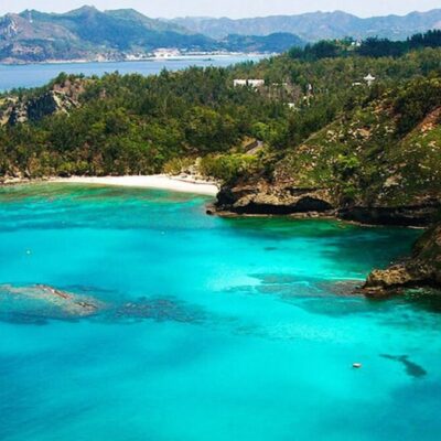 Stunning turquoise bay surrounded by lush green hills and rocky cliffs, with mountains visible in the distance