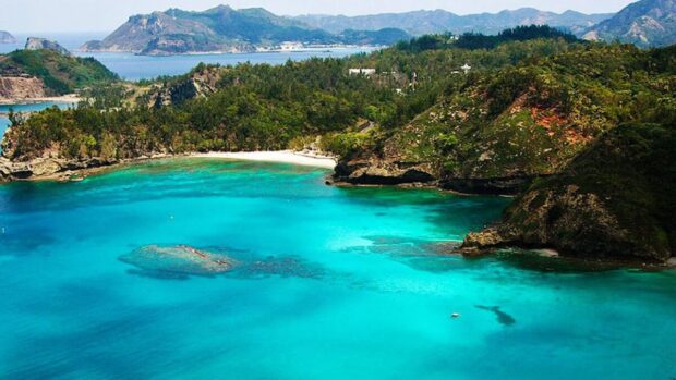 Stunning turquoise bay surrounded by lush green hills and rocky cliffs, with mountains visible in the distance