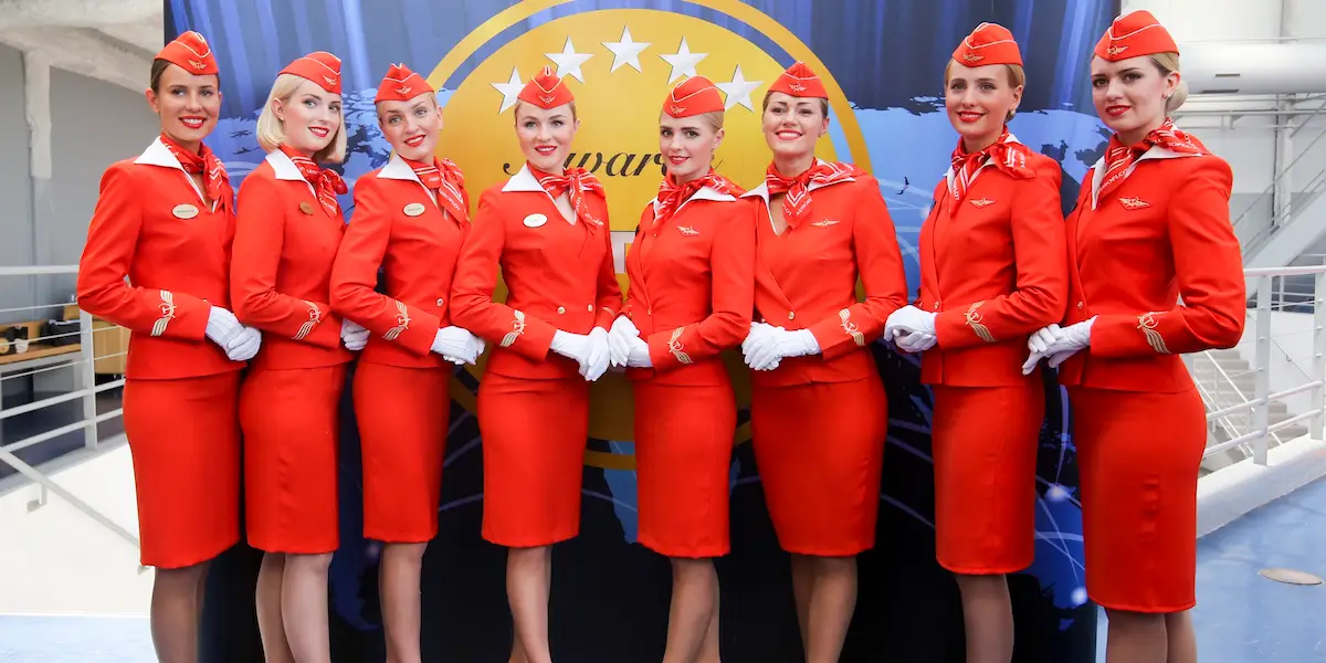 Eight flight attendants in matching red uniforms and hats pose together in front of an airline logo backdrop