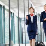Two professional flight attendants in navy uniforms with red scarves walking through modern airport terminal with luggage