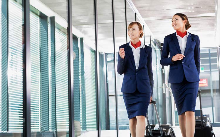 Two professional flight attendants in navy uniforms with red scarves walking through modern airport terminal with luggage